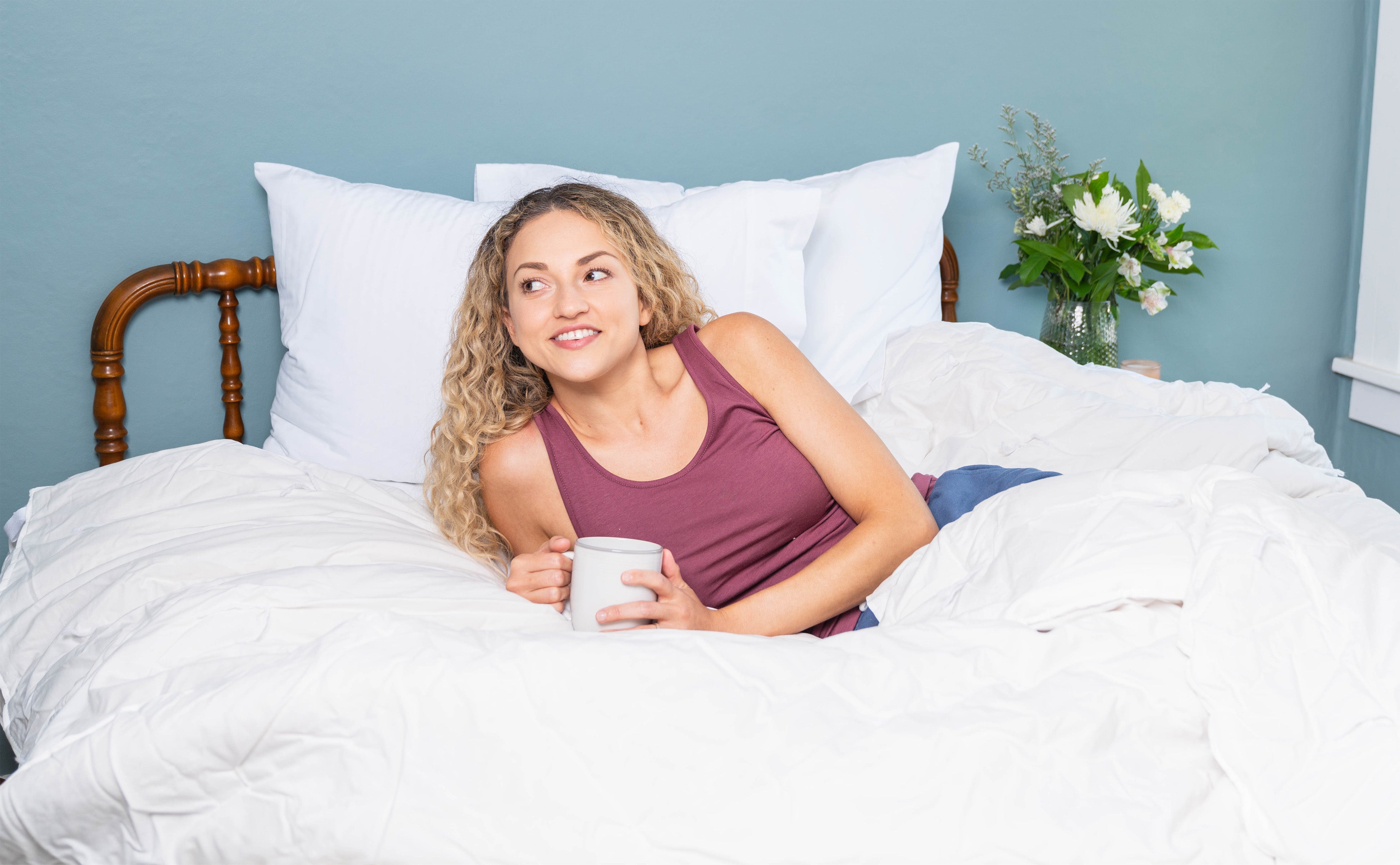 Woman lounging in bed (which is dressed with an A50 Duvet Cover, Custom Comfort Pillows, and Heritage American Sheets) enjoying a cup of coffee.