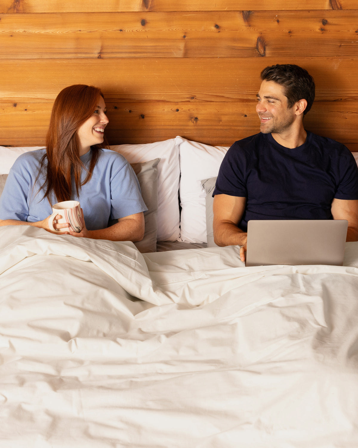 Couple sitting in bed, made with all Heritage American Sheets colors, but mainly Rustic Cotton, with a laptop and mug.