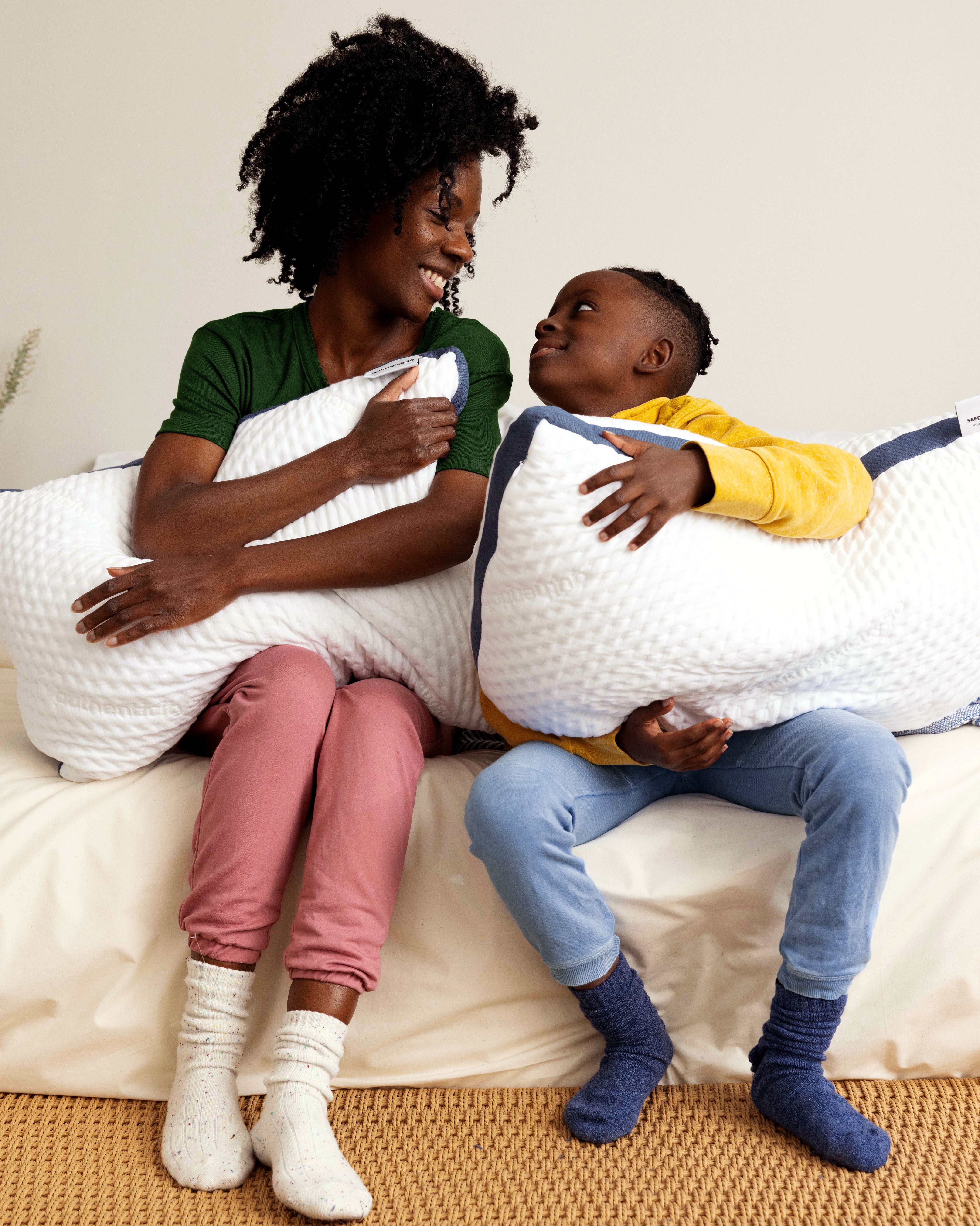 Mother and son smiling and hugging their Custom Comfort Pillows sitting on the edge of a bed.