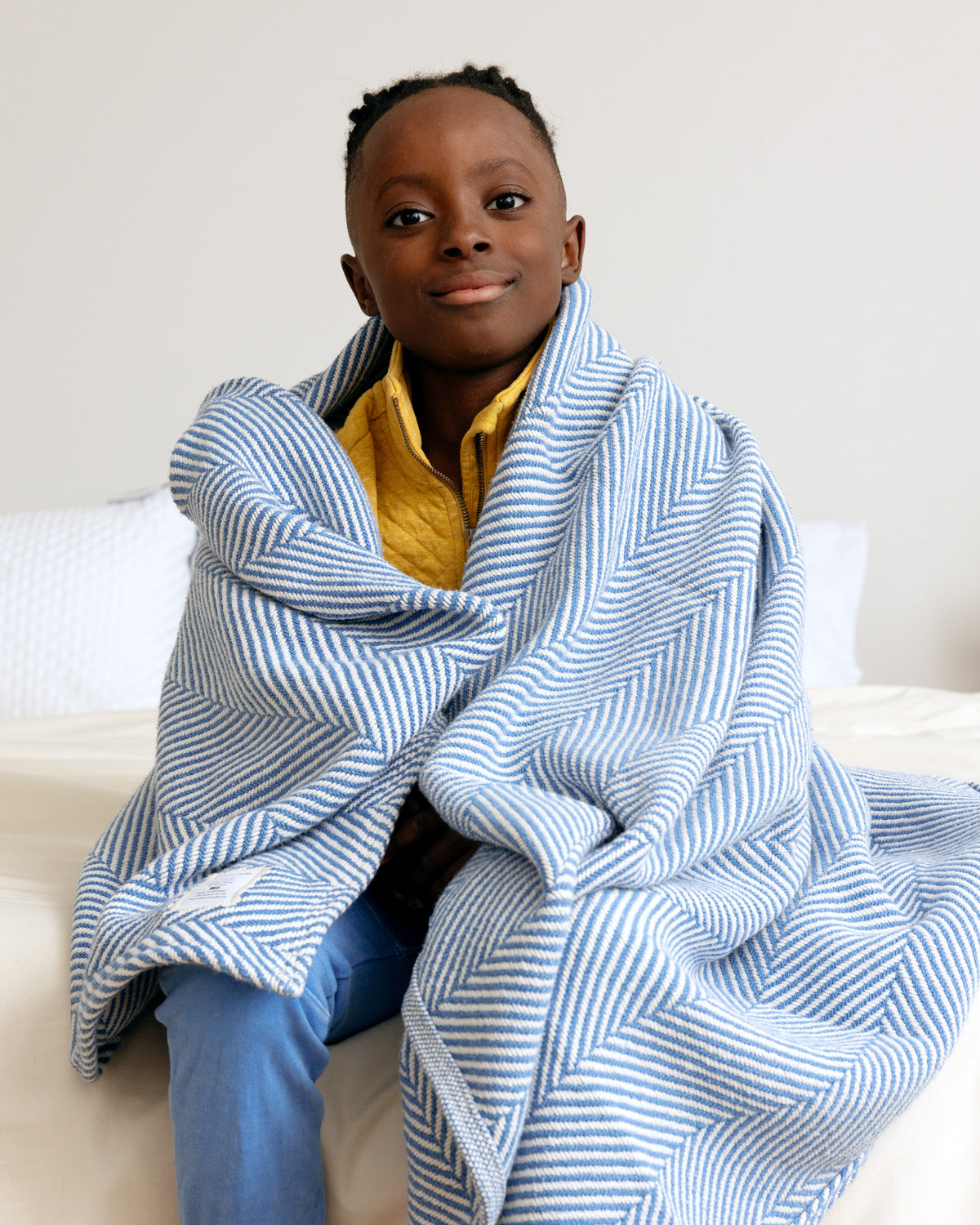 A boy sitting on the edge of a bed snuggled up with a Coastal Blue Heritage Blanket.