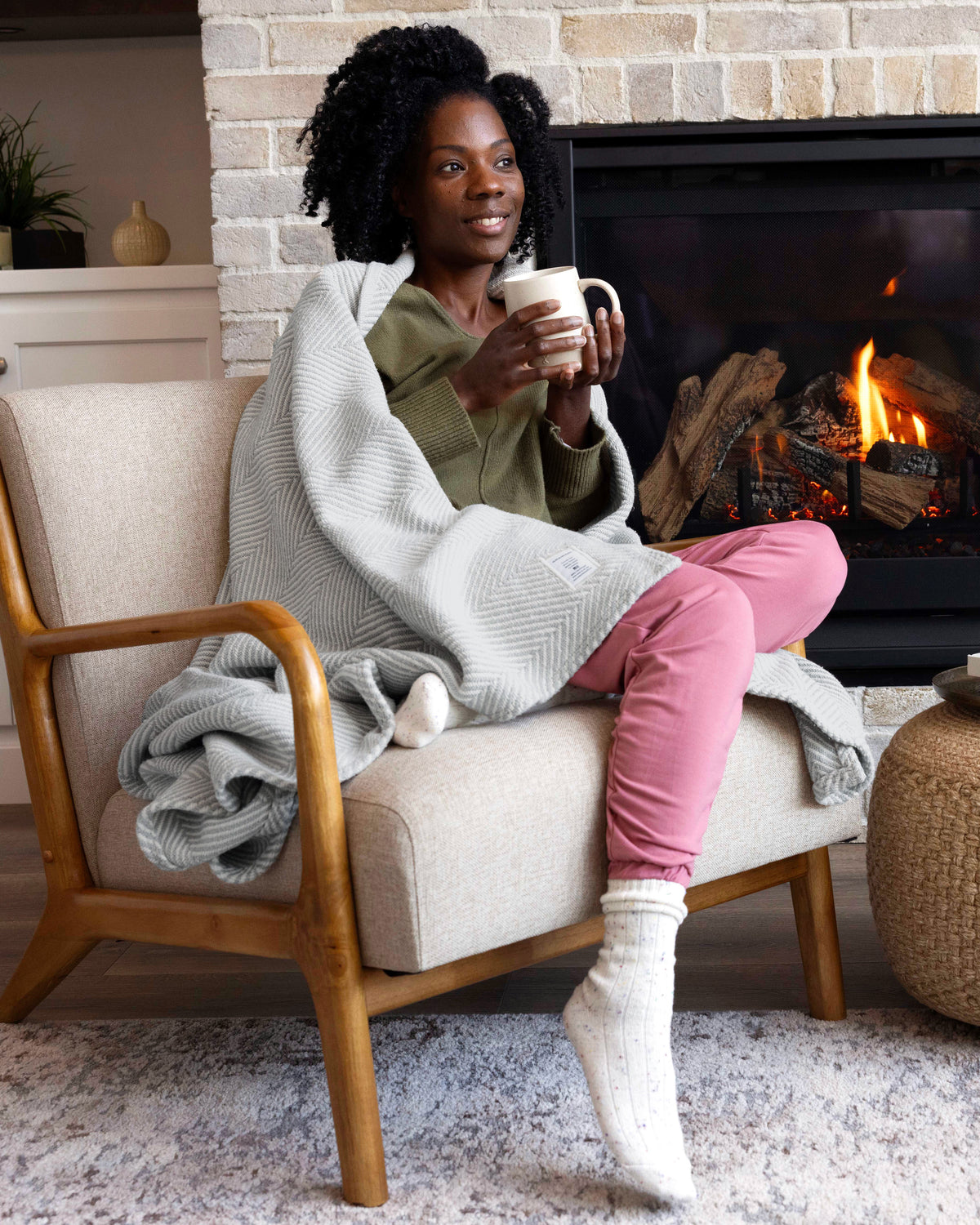 Woman relaxing by the fireplace with a mug and Cascade Gray Heritage Blanket.