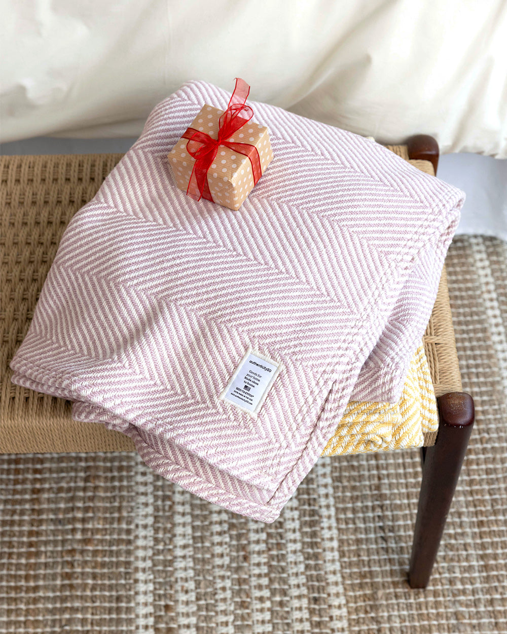 Two Heritage Blankets folded and stacked with a wrapped gift on a table at the foot of a bed.