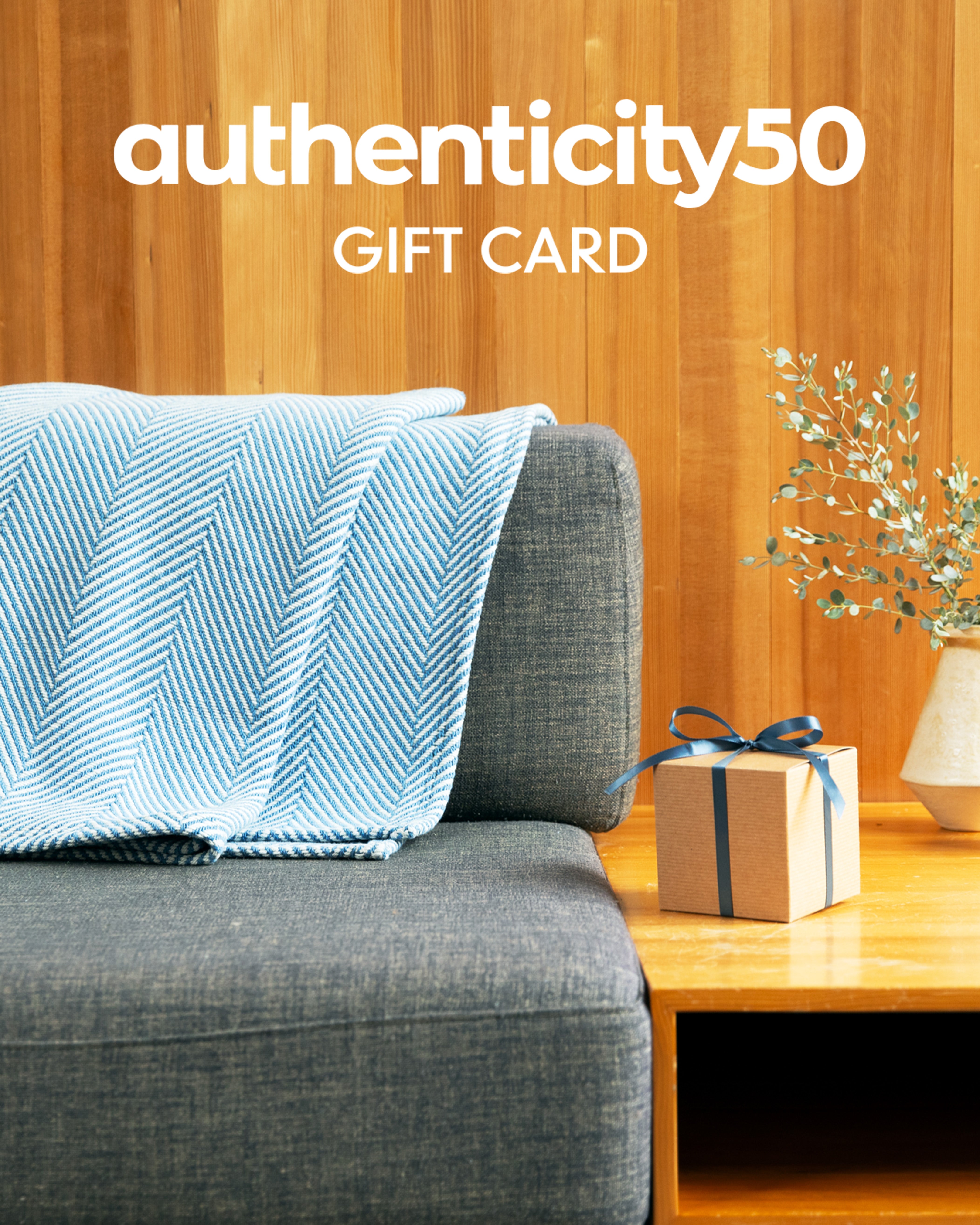 Folded Coastal Blue Heritage Blanket draped over the back of a couch next to a wrapped gift with the overlaying text: Authenticity50 Gift Card