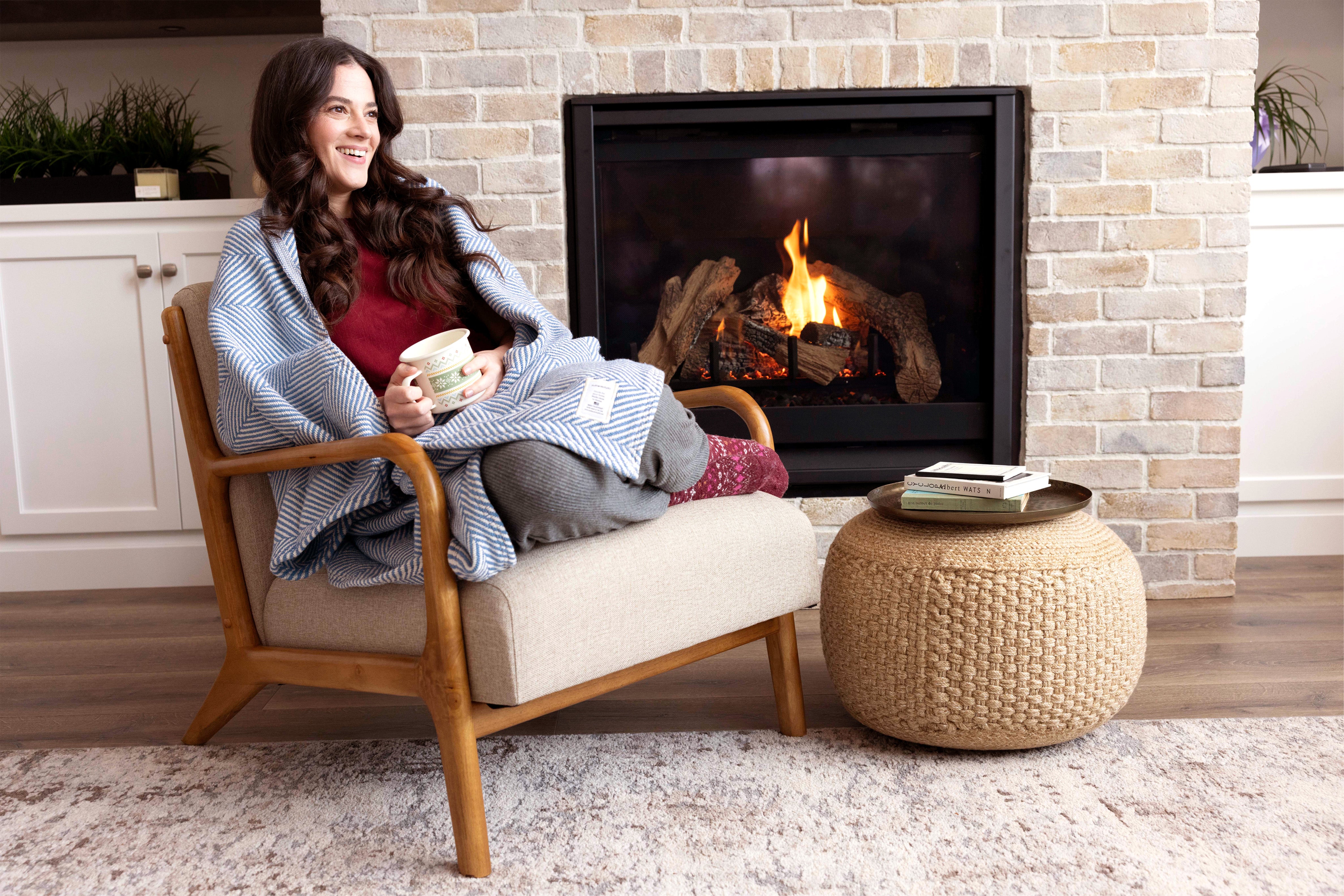 Woman relaxing by the fire with a cup of coffee and cozy Coastal Blue Heritage Blanket.