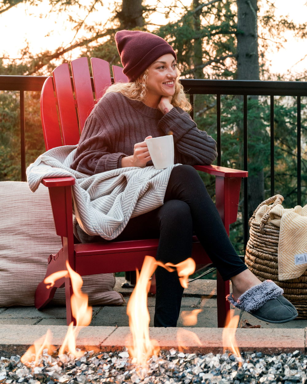 Woman sitting on her back deck by a fire pit with a Heritage Blanket on her lap and a mug in her hand.