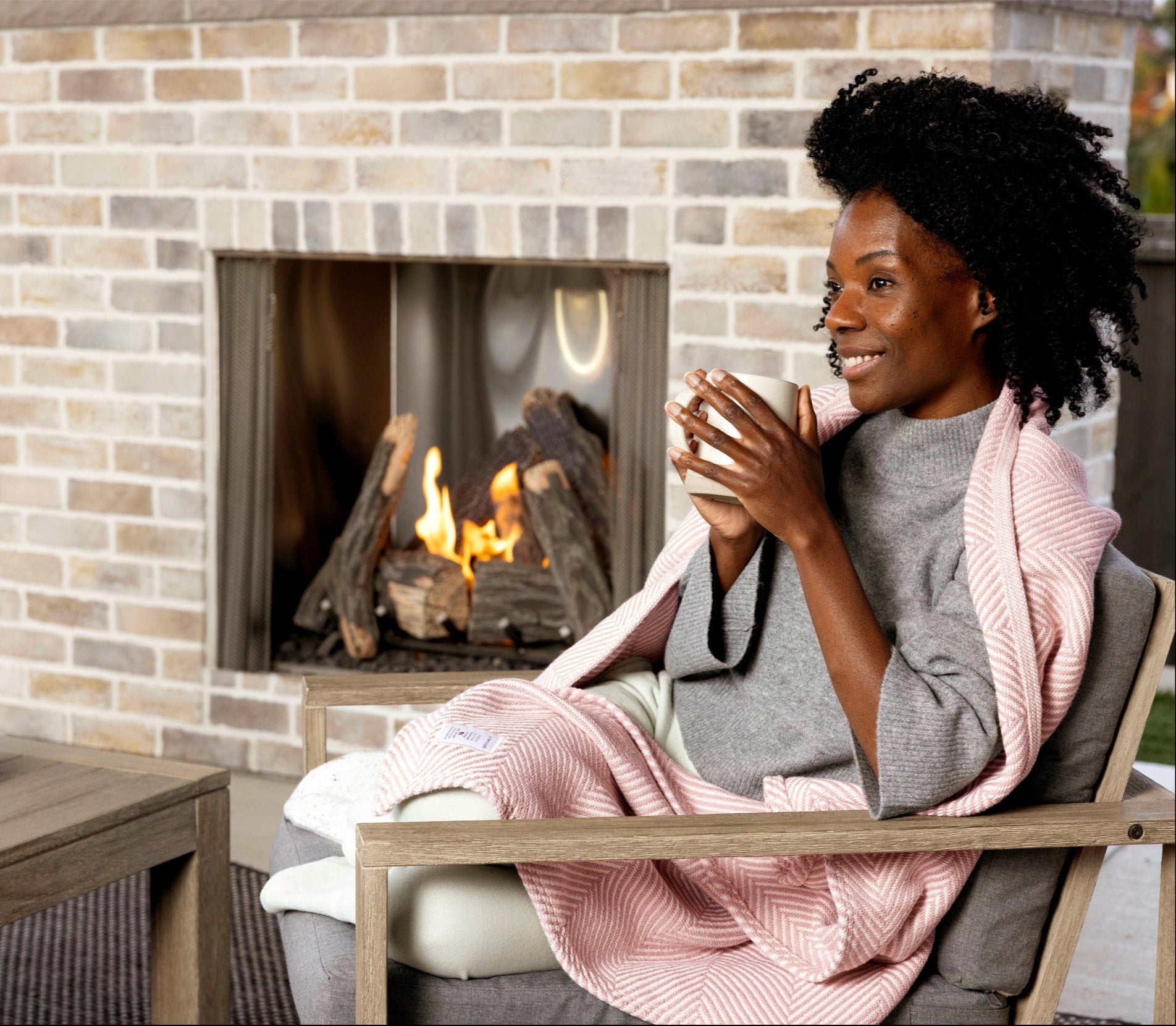 Woman sitting on her back porch by a fireplace smiling and holding a mug. She has a Desert Blush Heritage Blanket on her lap.