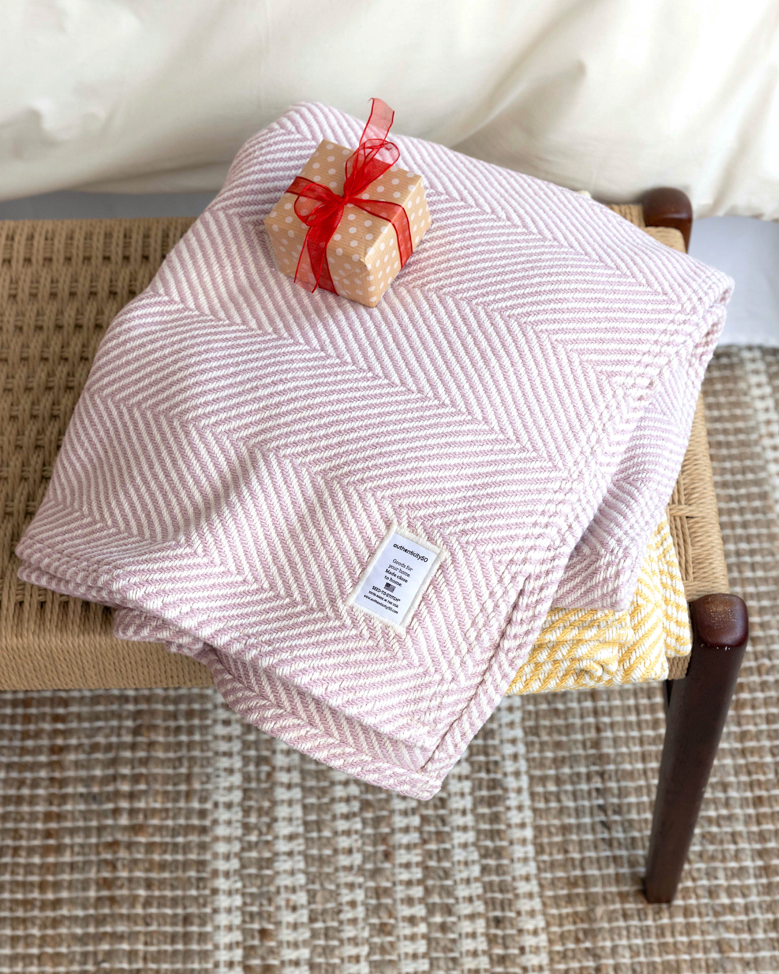 Stack of Heritage Blankets and wrapped gift stacked on a table a the foot of a bed.