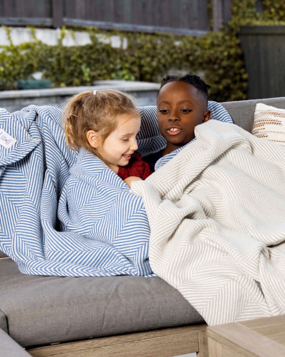 Two kids snuggled under their Heritage Blankets on a back patio.