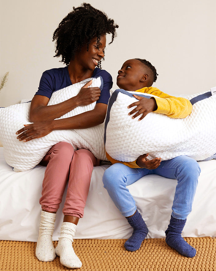 Mother and son sitting on a bed (which is made with Heritage American Sheets) hugging their Custom Comfort Pillows.