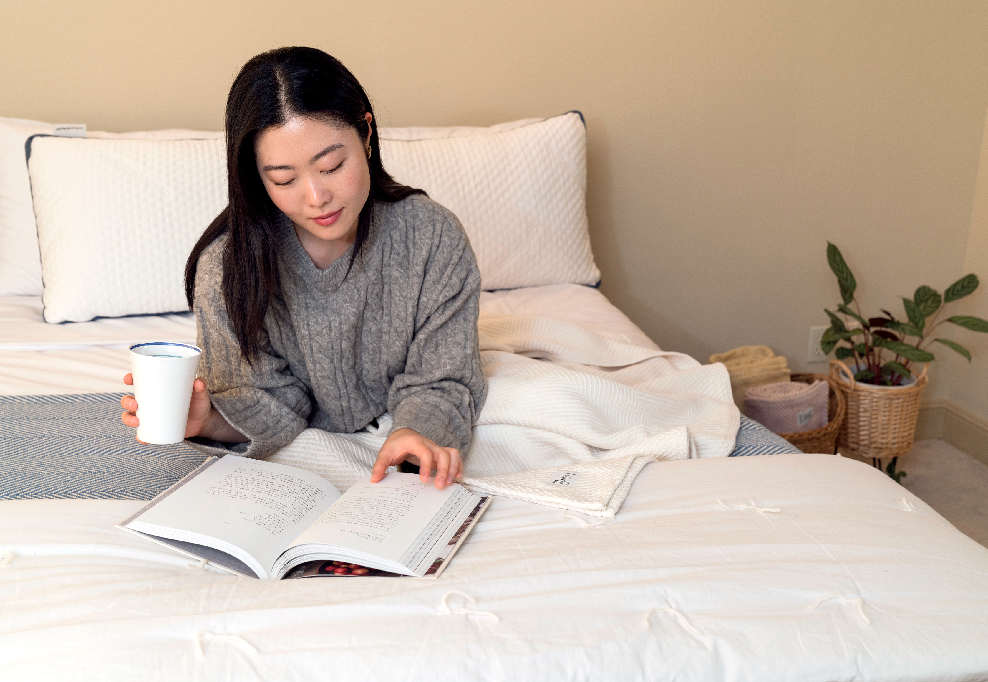 Woman reading in bed with a cup of coffee. The bed is made up with Custom Comfort Pillows, Heritage Blankets, and Heritage American Sheets.