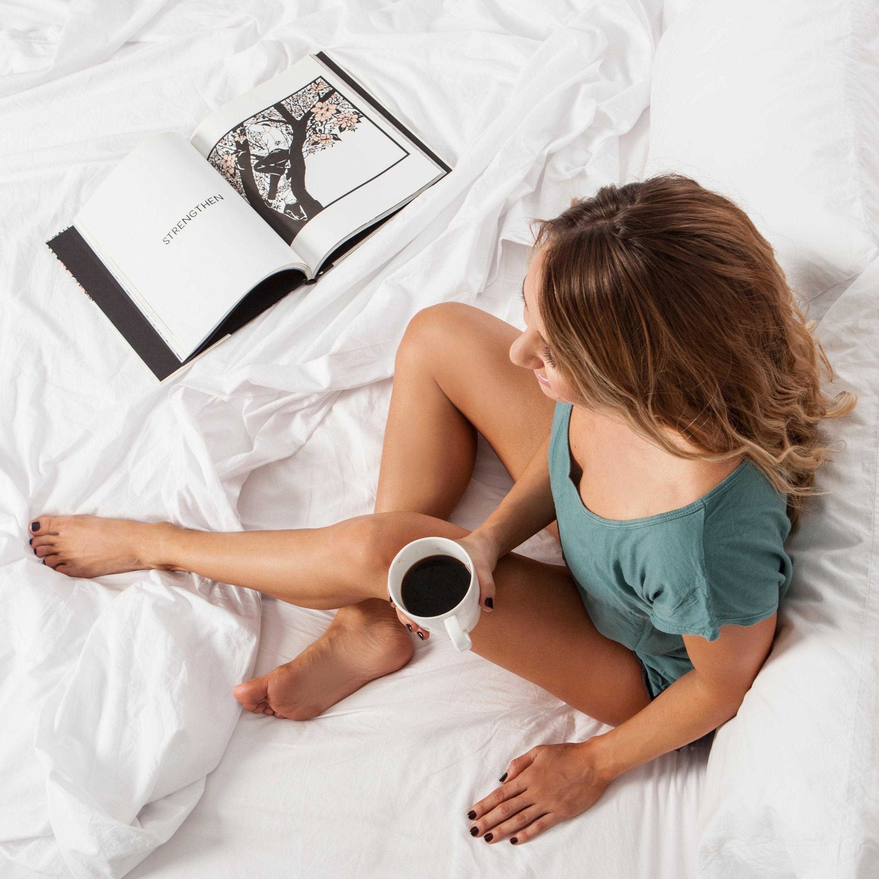 Woman sitting on a bed made of Cloud White Heritage American Sheets. She's holding a coffee and reading a book.